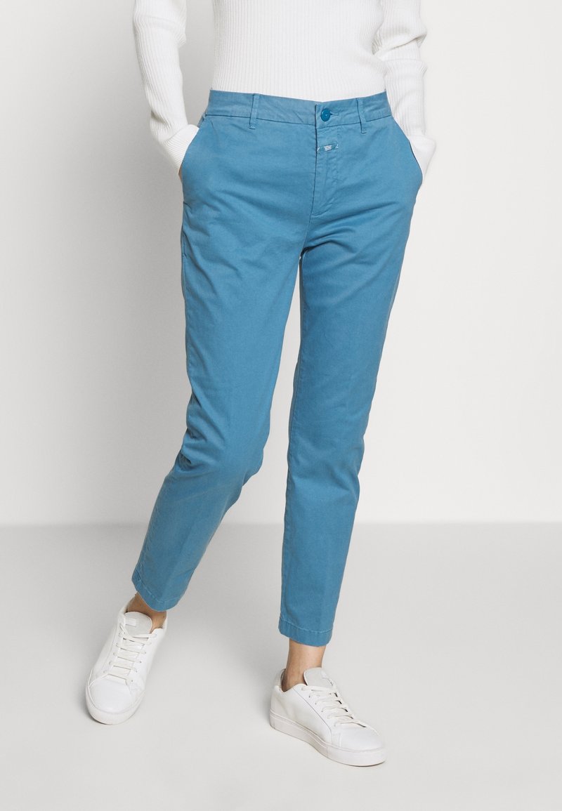 Pantaloni in cotone azzurro chiaro con vestibilità slim, dotati di tasche frontali, chiusura con bottone e caviglie regolabili, abbinati a scarpe da ginnastica bianche.