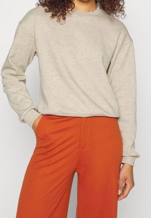 Persoon met krullend haar, gekleed in een beige shirt met lange mouwen en oranje broek, met één hand in de zak, tegen een effen lichtgekleurde achtergrond.