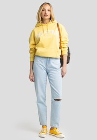 Sudadera amarilla con el logo "BIG STAR JEANS", jeans claros desgastados y zapatillas amarillas. Presenta una mochila marrón con correas.