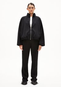 Frau steht in einer schwarzen, übergroßen Zip-Jacke, schwarzen geraden Hosen und schwarzen, fransigen Schuhen vor einem schlichten weißen Hintergrund.