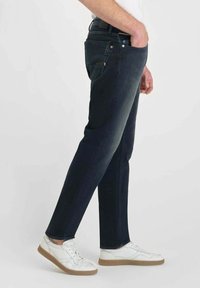 Jean en denim bleu foncé avec une coupe slim, présentant des poches latérales, une fermeture à bouton et une texture lisse, associés à des baskets blanches.