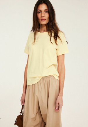 Kobieta z długimi ciemnymi włosami nosi jasny żółty t-shirt, beżowe spodnie z szerokimi nogawkami i trzyma brązową torbę z sznurkiem przy boku.