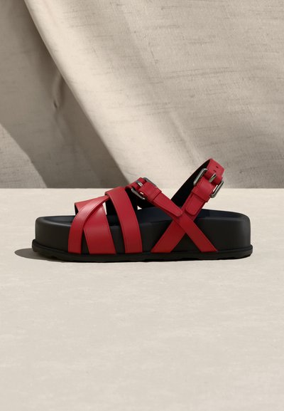 Sandale en cuir rouge avec semelle plateforme noire, brides ajustables à boucle, et design croisé, présentée sur une surface beige avec un fond en tissu.