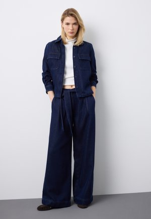 Donna bionda in piedi contro un muro bianco, indossa una giacca di jeans blu scuro e pantaloni in denim a gamba larga abbinati a un top corto bianco e scarpe marroni.