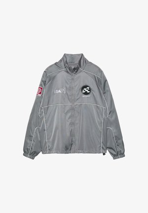 Siva športna jopa z zadrgo, belimi obrobami, napisom "USA 91" na prsih, črnim krožnim športnim logotipom in rdečo značko na levi rokavu.