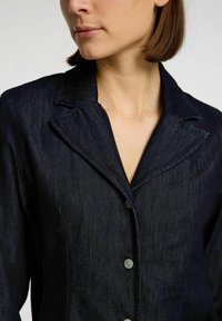 Blazer di denim navy con colletto a punta, bottoni frontali e una finitura texturizzata. Il design include cuciture sartoriali e una forma strutturata.