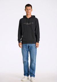 Sudadera negra con el texto "COPENHAGUE", mangas largas y un bolsillo frontal; combinada con vaqueros azul claro y zapatillas blancas.