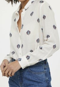 Chemise blanche à manches longues avec col, ornée d'une broderie colorée en forme de diamant en bleu, marron et noir. Accordée avec un jean en denim bleu.