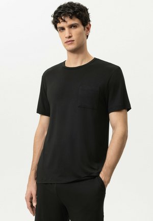 Junger Mann mit lockigem Haar, der ein schlichtes schwarzes Kurzarm-T-Shirt mit Brusttasche trägt und mit einer Hand in der Hosentasche steht.