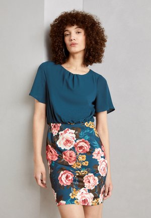 Türkise Bluse mit gerafftem Ausschnitt, kombiniert mit einem figurbetonten Rock mit Blumenmuster, der pinke Rosen und gelbe Akzente auf blauem Hintergrund zeigt.