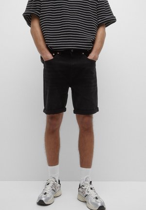Persona con pantalones cortos negros remangados, camisa de rayas blancas y negras, calcetines blancos y zapatillas de correr plateadas, de pie con las manos en los bolsillos.