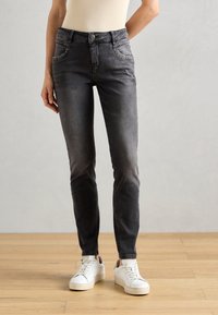 Szürke farmernadrág, slim fit fazonban, fém szegecsekkel a zsebeken, fehér sportcipőkkel, fából készült padlón.