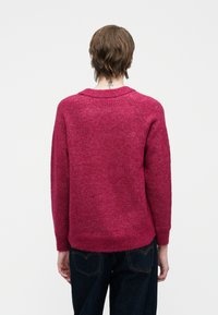 Osoba z krótkimi brązowymi włosami, ubrana w czerwoną bluzę i ciemne dżinsy, zwrócona plecami, z małym tatuażem na karku.