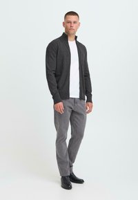 Hombre de pie, vistiendo un suéter con cremallera gris oscuro sobre una camiseta blanca, jeans grises y zapatos negros, contra un fondo claro y liso.