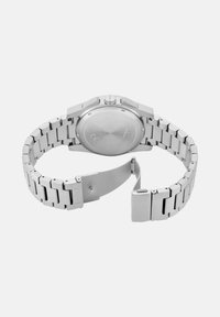 Srebrna kovinska ura z zapestnico iz nerjavečega jekla, okroglo ohišje in gladko, trdo zadnjo stran. Ima sponko za zapiranje.