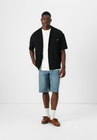 Schwarzes kurzärmliges Hemd über einem weißen T-Shirt, kombiniert mit blauen Jeansshorts und braunen Bootsschuhen. Flache Textur mit dezentem Branding-Detail.