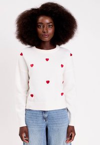 Femme avec de grandes boucles de cheveux, portant un pull blanc à petits cœurs rouges dispersés et un jean bleu clair, faisant face à l'avant contre un fond blanc.