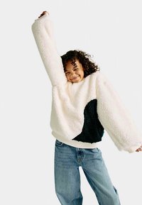 White fleece sweatshirt with a black textured accent, wide sleeves, and neckline. Paired with light blue denim jeans.