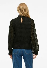 Blusa nera in pizzo con maniche lunghe e gonfie, scollatura alta e apertura a chiave sul retro; abbinata a jeans blu a vita alta.