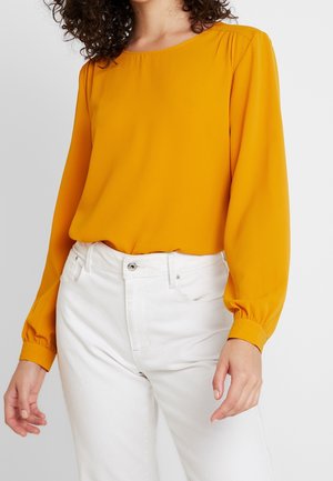 Kobieta w musztardowożółtej bluzce z długim rękawem wpuszczonej w spodnie z wysokim stanem w kolorze białym, stojąca na jednolitym tle.