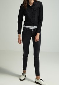 Chaqueta de mezclilla negra con un diseño ajustado y cierre de botones, combinada con jeans skinny negros. Unos zapatos blancos con cordones completan el conjunto.