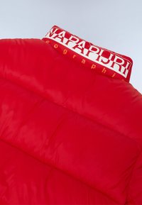 Rød polstret jakke med høj krave, der har et hvidt logo og tekst. Det glatte, blanke stof har fremtrædende syninger og quiltede sektioner.