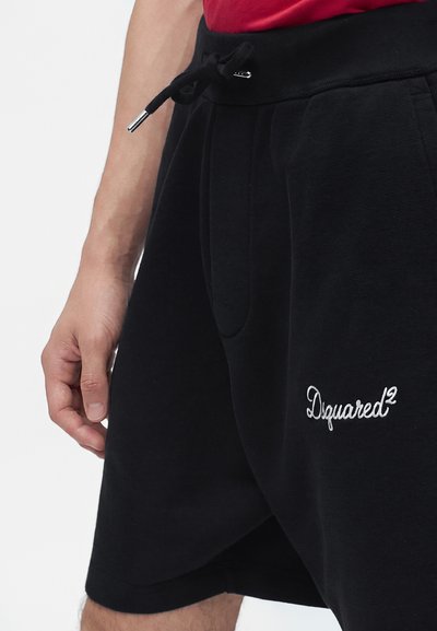 Gros plan d'une personne portant un short noir avec une taille à cordon et un logo blanc "Dsquared2" sur la jambe gauche.