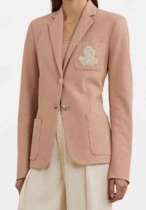 Femme portant un blazer texturé rose clair avec des boutons argentés et un emblème brodé sur la poche poitrine, associé à un pantalon couleur crème.