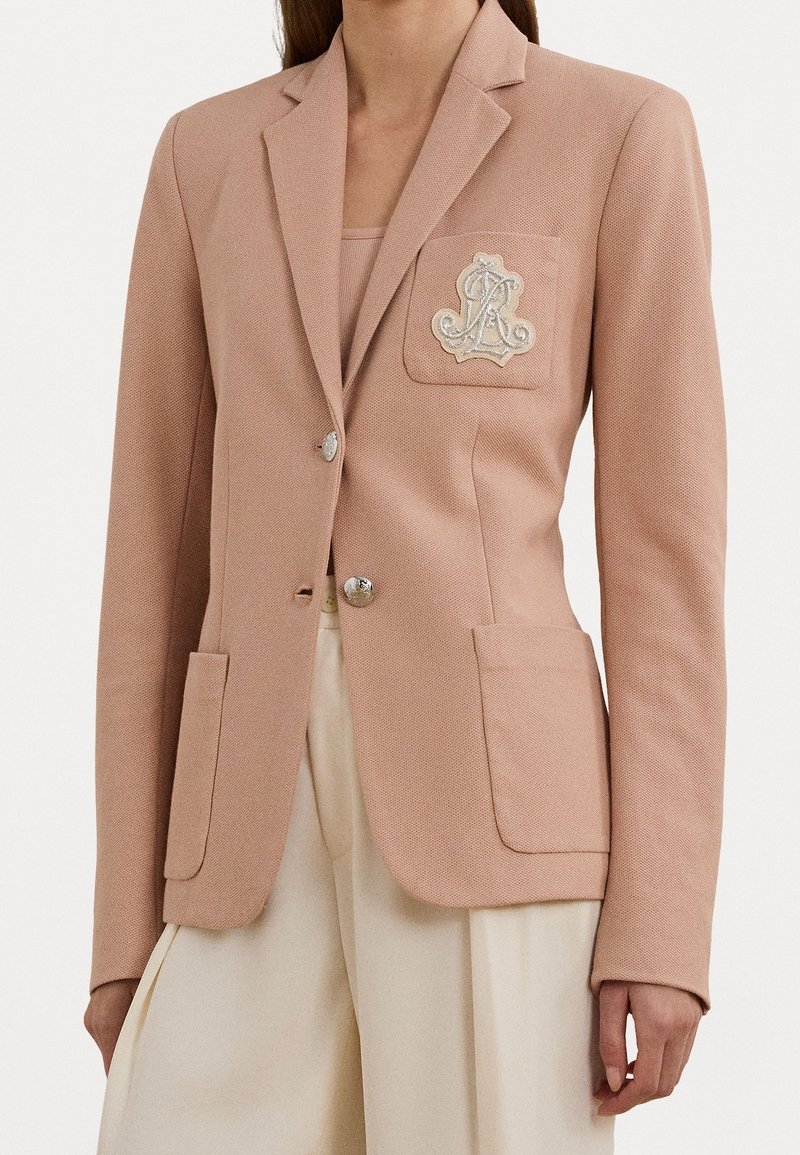 Femme portant un blazer texturé rose clair avec des boutons argentés et un emblème brodé sur la poche poitrine, associé à un pantalon couleur crème.