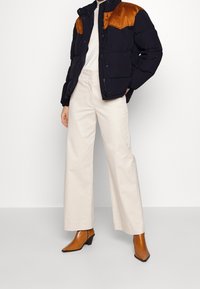 Puchowa kurtka w granatowym kolorze z brązowymi zamszowymi akcentami na ramionach, zestawiona z szerokimi białymi spodniami i brązowymi botkami na obcasie. Minimalistyczny design.