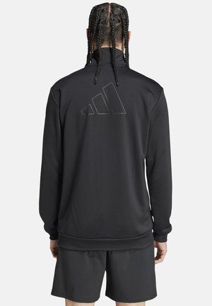 Person mit geflochtenem Haar trägt eine schwarze Langarmjacke mit einem dezenten Adidas-Logo auf dem Rücken und schwarze Shorts, von hinten gesehen.