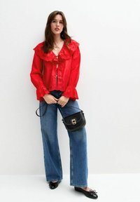 Blouse rouge avec un col froncé et des détails en œillets, portée avec un jean large bleu et un sac bandoulière noir. Des ballerines noires complètent le look.