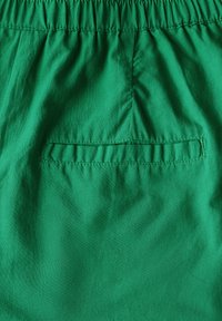 Zelené šortky s elastickým pasem a dekorativní zadní kapsou. Materiál je hladký s mírným leskem, což naznačuje lehkou tkaninu.