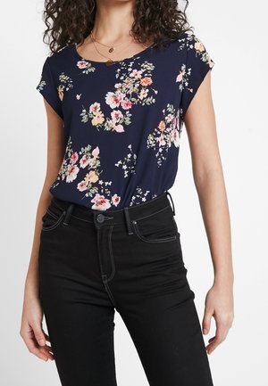 Vrouw draagt een marineblauw bloesje met korte mouwen met bloemenprint, ingestopt in een zwarte high-waisted spijkerbroek met zichtbare stiksels en een knoopsluiting.