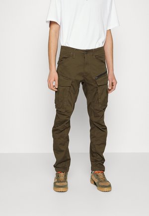 Hombre con pantalones cargo verde oliva con múltiples bolsillos y cremalleras, combinado con una camiseta blanca y zapatillas de camuflaje.