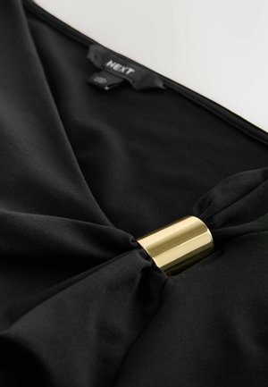 Zwarte kledingstuk met een gouden cilindervormige sluiting die de stof bij de hals bijeenbrengt, met een klein Next-merkje aan de binnenkant.