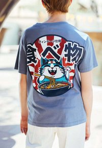 Enfant portant un t-shirt bleu avec un dessin d'un chat de dessin animé souriant mangeant des nouilles avec des baguettes, accompagné de texte japonais au dos.