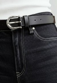 Zwarte leren riem met een zilveren metalen gesp, gedragen over donkerdenim jeans met contrasterende witte stiksels en een fitted witte blouse.