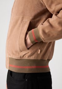 Tansue Bomberjacke aus Wildleder mit gerippten, olivgrünen Bündchen, die einen roten Streifen aufweisen. Seitentaschendetails sichtbar, kombiniert mit schwarzen Hosen.