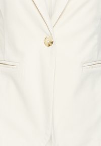 Blazer crème avec un seul bouton marbré, un revers cranté et deux poches fendues horizontales à l'avant.