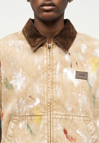 Κοντινό πλάνο ατόμου που φοράει μπεζ μπουφάν με πιτσιλιές μπογιάς, καφέ γιακά από κοτλέ και ετικέτα Moschino στο στήθος, με το φερμουάρ κλειστό μπροστά.