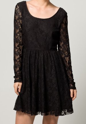 Vestido negro de encaje con mangas largas y escote en forma de barco, que presenta un patrón floral y una falda acampanada. La tela texturizada añade elegancia.