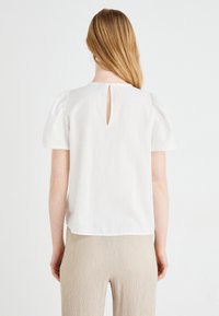 Femme aux longs cheveux châtain clair portant un chemisier blanc à manches courtes avec une ouverture en forme de trou de serrure dans le dos et un pantalon beige texturé, debout sur fond blanc.