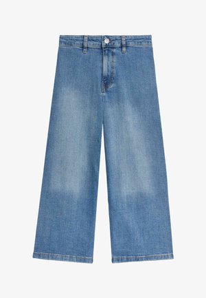 Biała bluza z polaru z kieszenią z przodu, zestawiona z szerokimi niebieskimi dżinsami. Model nosi beżowe sandały z klamrami.