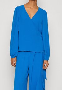 Blue wrap blouse with long balloon sleeves, textured fabric, and a deep V-neckline, paired with matching wide-leg trousers.