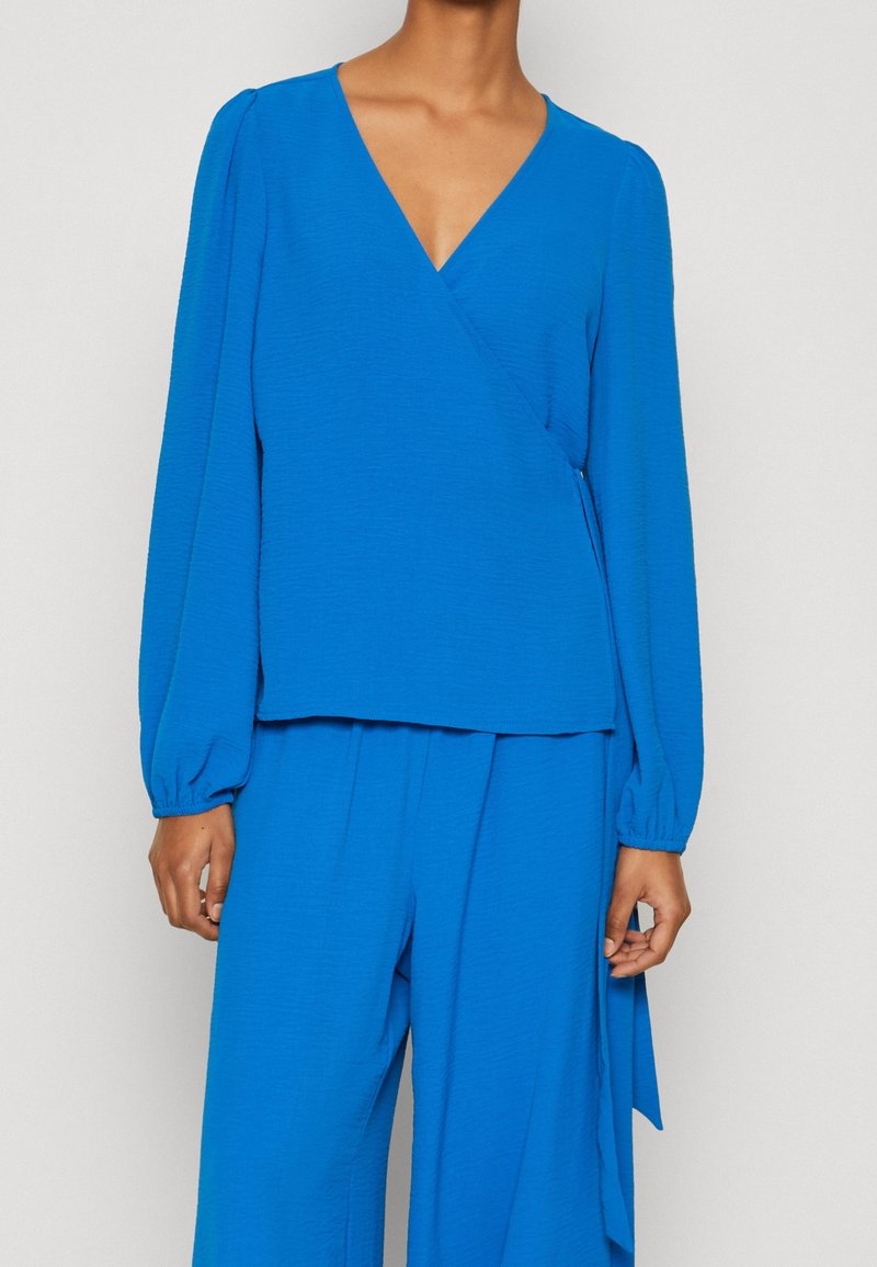 Blue wrap blouse with long balloon sleeves, textured fabric, and a deep V-neckline, paired with matching wide-leg trousers.