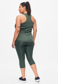 Groene sportieve tanktop en capri legging met een gladde textuur. Heeft een racerback ontwerp en een enkel-lange snit, gecombineerd met zwarte schoenen.