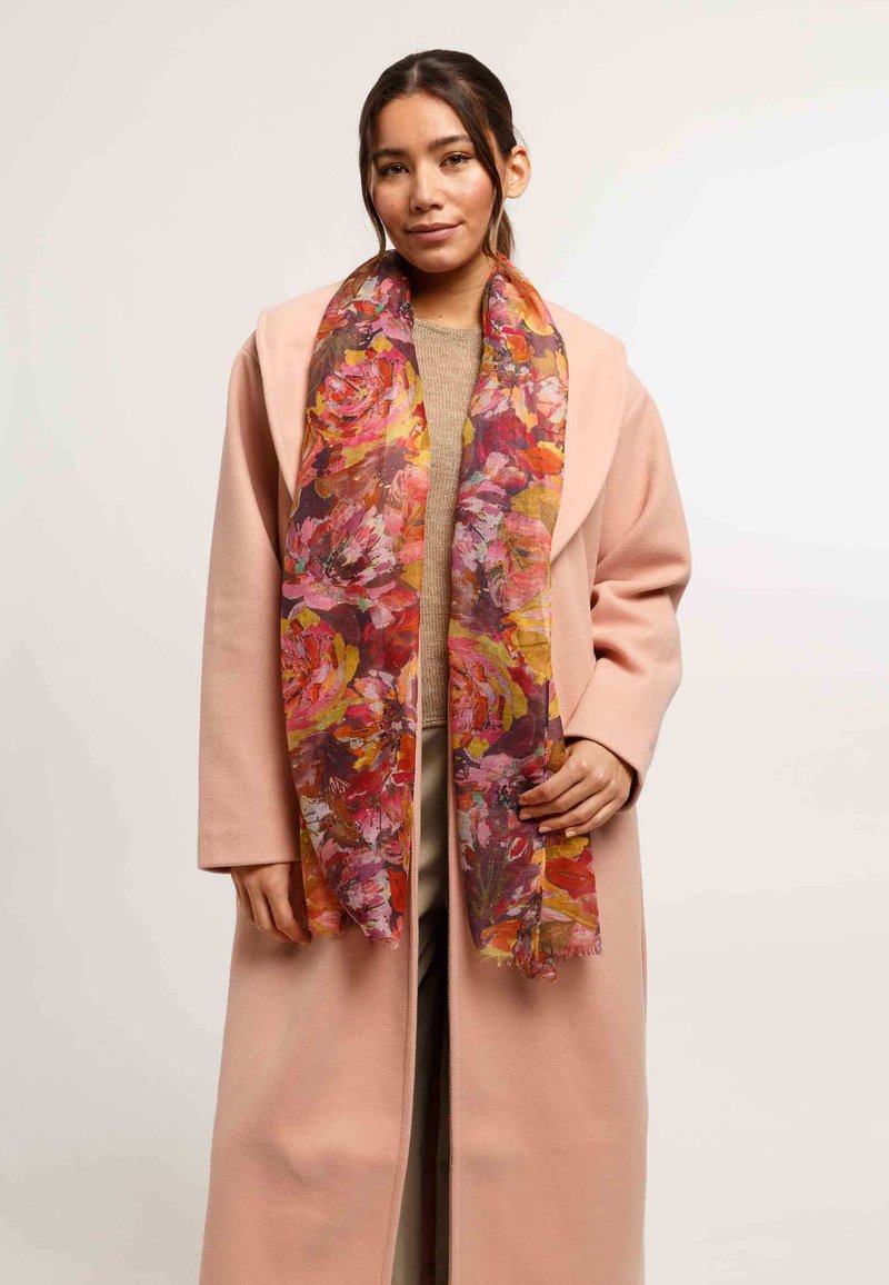 Scarf floreale leggero con motivi vibranti in rosa, arancio e giallo. Texture trasparente che si drappeggia elegantemente. Indossato sopra un maglione beige e un cappotto rosa.