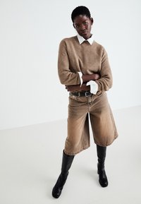 Pull en maille marron avec une chemise blanche en dessous, associé à des culottes en denim beige et des bottes noires jusqu'au genou.