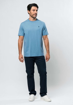 Vīrietis, valkājot gaiši zilu īsroku krekls, tumšas džinsa bikses un baltas sporta kurpes, stāv priekšā vienkāršai gaiši pelēkai fona sienai.